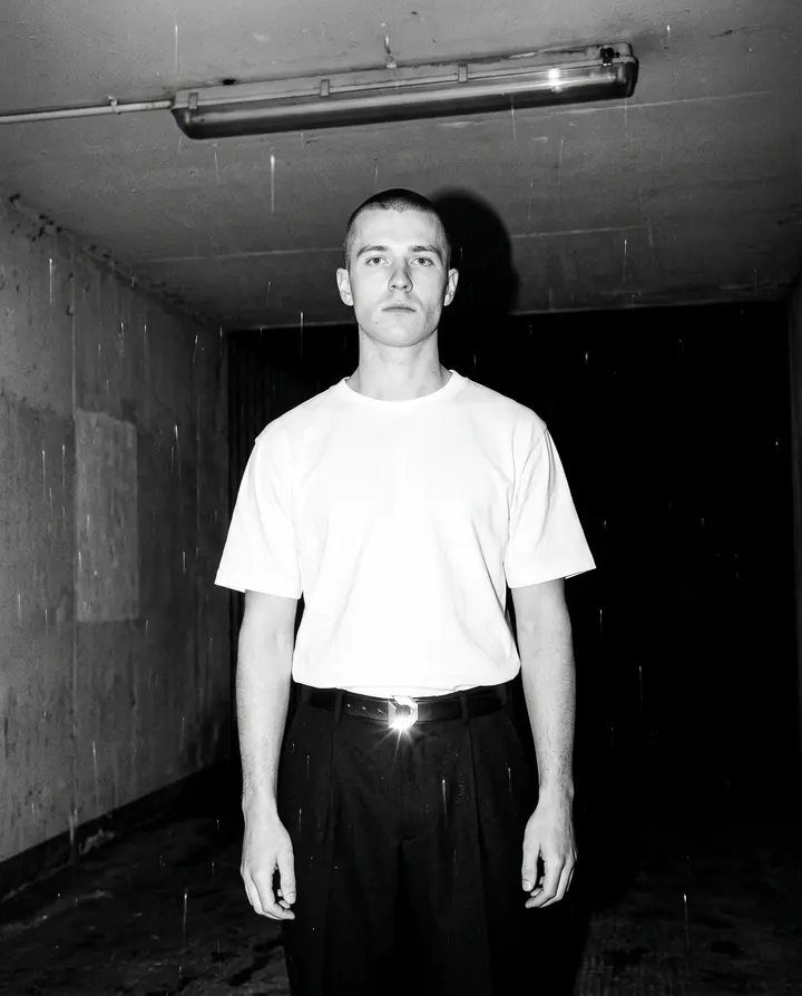 A man in a rain-wet underpass in the white Heavyweight Tee and Pleated Trouser, the steel buckle of the Dress Belt flaring in the flash.